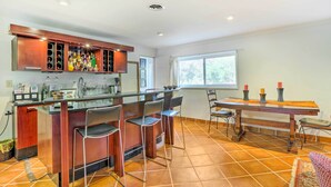 A kitchen with a bar area, bar stools, a dining table with chairs, and a window with curtains.
