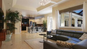 A spacious living room with a black leather sofa, a coffee table, and a chandelier. There is a kitchen in the background with a bar area. - Country Club Residence by 123cape VR (Fort Myers)