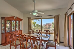 Un coin repas avec une table et des chaises en bois, un buffet avec des portes en verre, et une vue sur les palmiers et les montagnes à travers de grandes fenêtres.