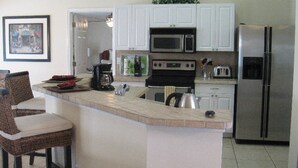 A kitchen with white cabinets, a stainless steel refrigerator, a microwave, and a stove. There is a bar area with wicker chairs. - Yvonne by 123cape VR (Cape Coral)
