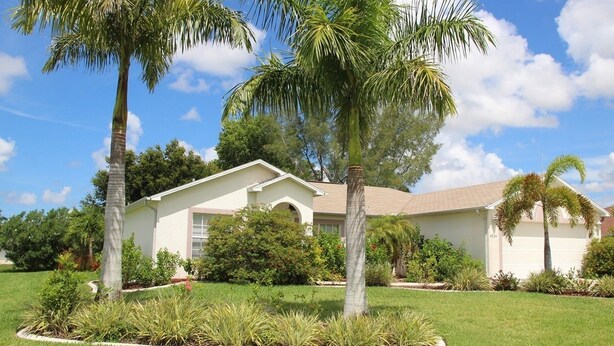 A single-story house with a white exterior, a garage, and a well-maintained lawn with palm trees and shrubs. - Yvonne by 123cape VR (Cape Coral)