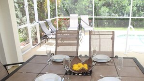 A sunlit patio with a table set for four, surrounded by chairs, and a view of a swimming pool and greenery. - Yvonne by 123cape VR (Cape Coral)