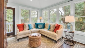 Sala de estar con sofá seccional color beige, un reposapiés tejido y una mesita redonda con lámpara. Amplias ventanas ofrecen vistas a zonas verdes exteriores.