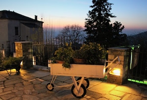A stone-paved patio with a wheelbarrow filled with plants, a stone wall, and a building in the background. - Archontiko Panagoula (Volos)