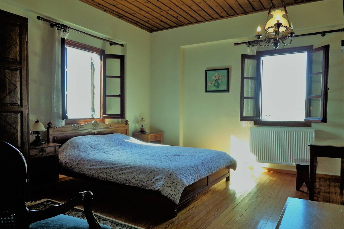 A bedroom with a wooden floor, a bed with a floral bedspread, a nightstand with a lamp, a chair, and a window with open shutters.