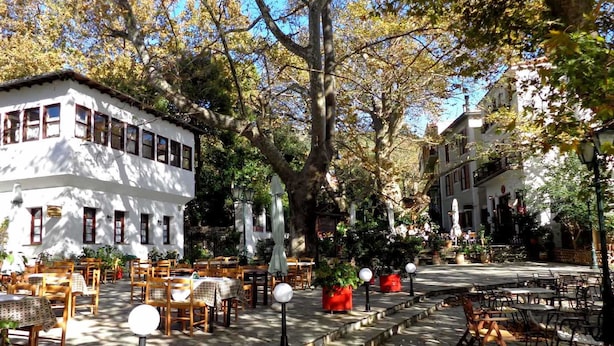An outdoor café with tables and chairs, surrounded by trees and buildings. - Archontiko Panagoula (Volos)
