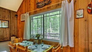 Ein Esszimmer mit Holzboden und einem großen Fenster mit Blick auf einen Wald, ein Holztisch gedeckt für vier Personen und ein dekoratives Schiff an der Wand.