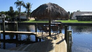 Un muelle de madera con una sombrilla de paja, sillas y una mesa junto a un cuerpo de agua tranquilo.