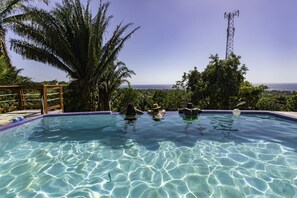 A clear blue pool with two people swimming, surrounded by lush greenery and a view of the ocean.