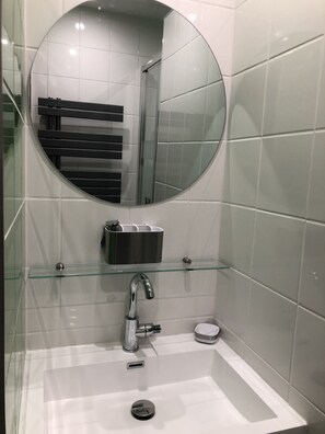Ett modernt badrum med en rund spegel, en hylla i glas med toalettartiklar, ett handfat med blandare och en toalett.