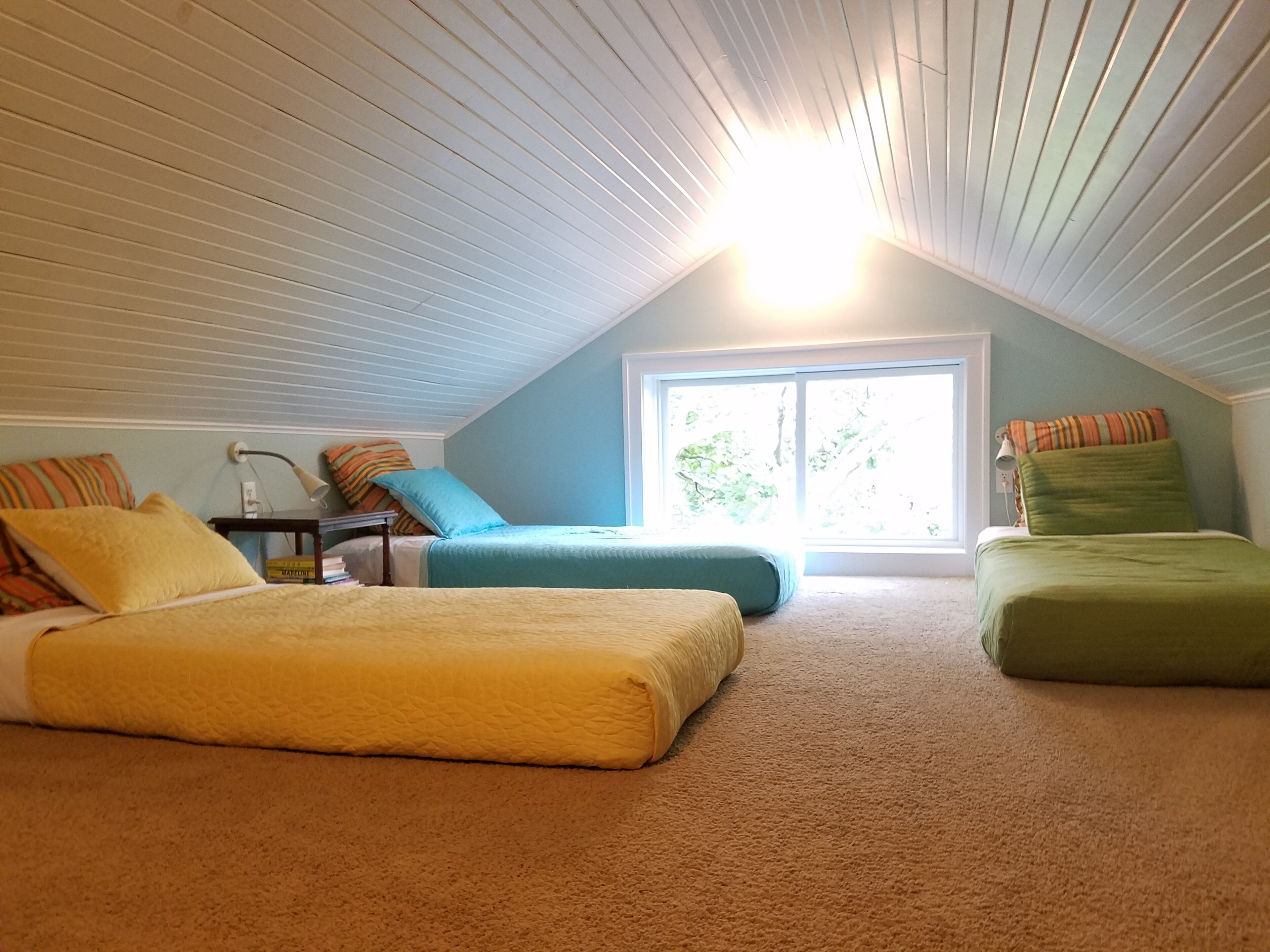 A cozy attic bedroom with two beds, a nightstand, and a window with a view of greenery.
