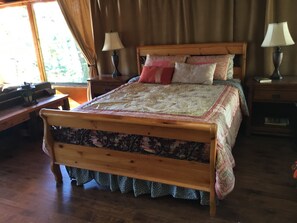 Una cama de madera con una colcha estampada, dos mesitas de noche con lámparas y una ventana con cortinas.