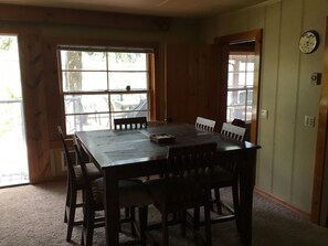Comedor con mesa y sillas de madera, ventanas amplias y un reloj en la pared.