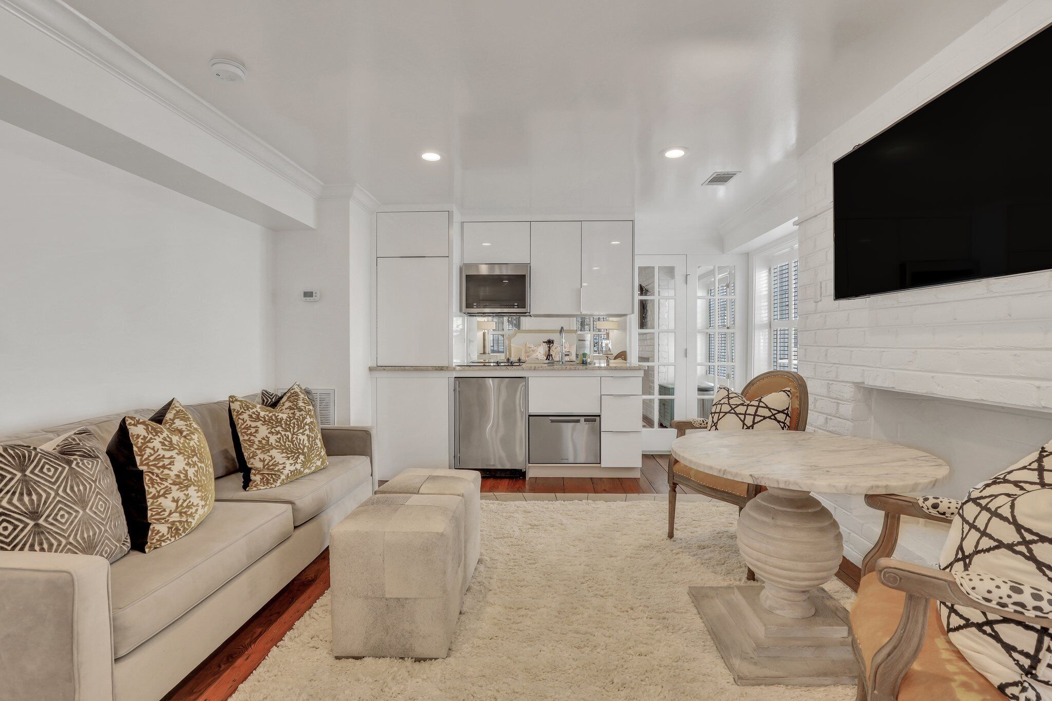 A modern living room with a sofa, ottoman, and a dining table with chairs. A kitchen with a stainless steel refrigerator and microwave is visible in the background.