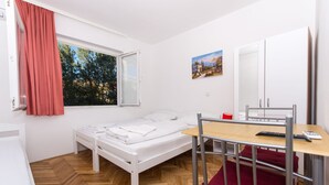 Une petite chambre à coucher, bien rangée, avec un lit simple, un bureau en bois, une chaise et une fenêtre donnant sur des arbres.