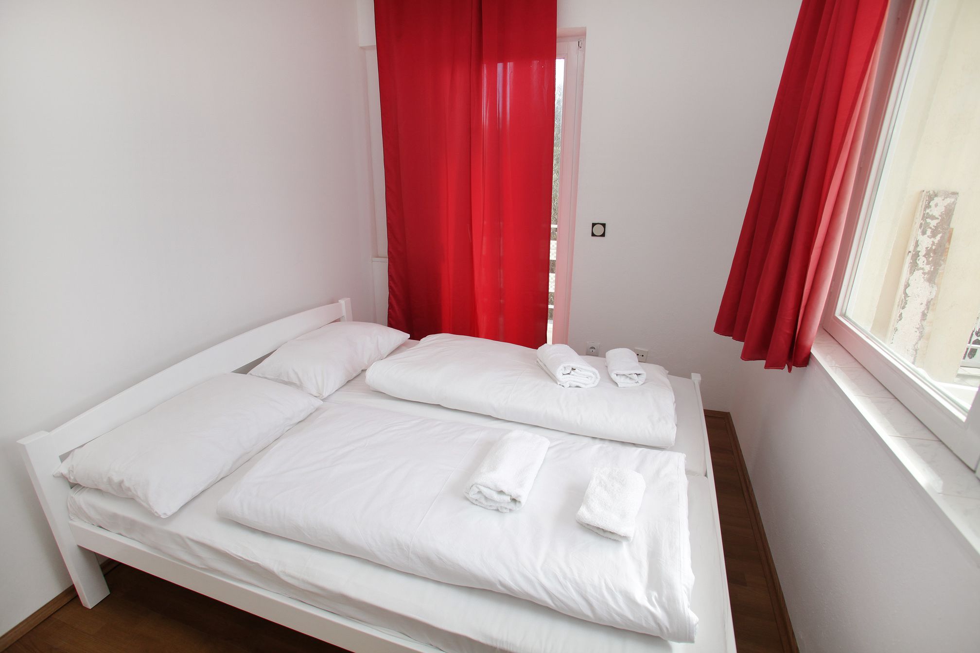 A single bed with white bedding and red curtains, a window with a view, and a small bedside table with a lamp.