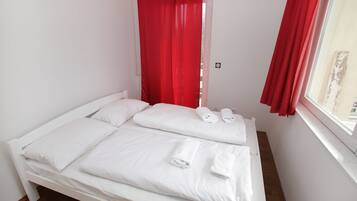 A single bed with white bedding and red curtains, a window with a view, and a small bedside table with a lamp.