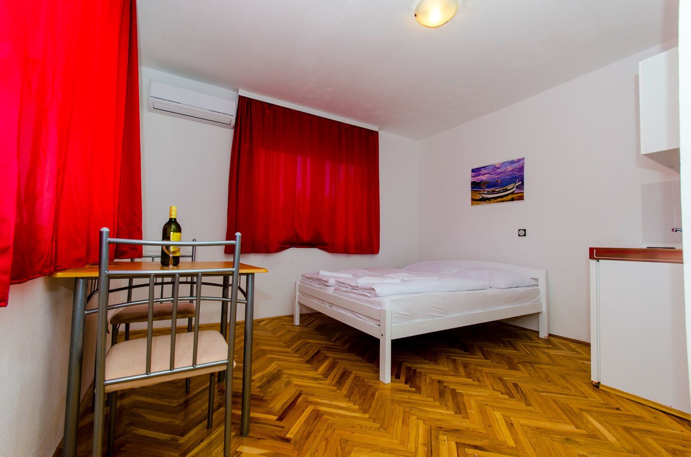 A small room with a bed, a table with a chair, a bottle, and a painting on the wall.