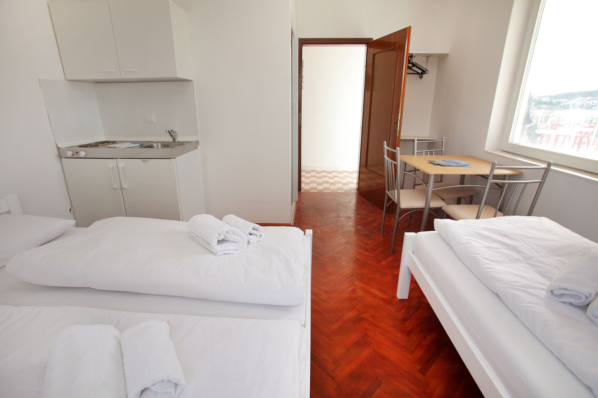 Una camera d'albergo con due letti, un angolo cottura, un'area pranzo con tavolo e sedie, e vista attraverso una porta aperta.