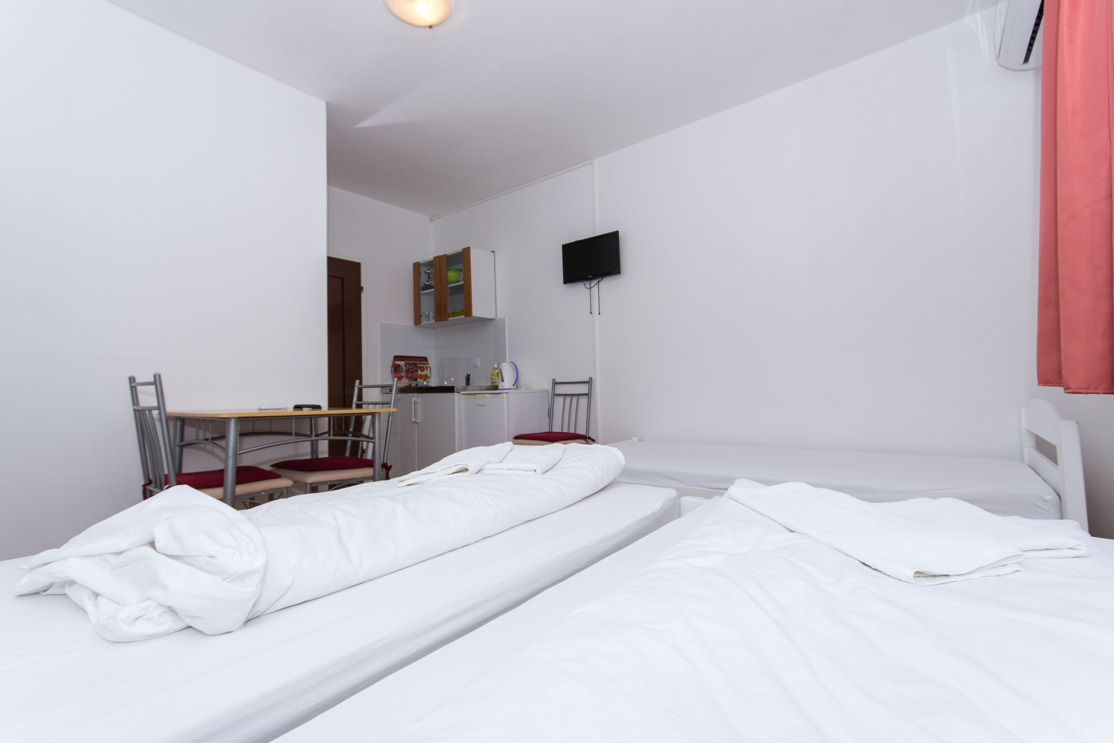 A room with two beds, a desk, a chair, a television, and a kitchenette.