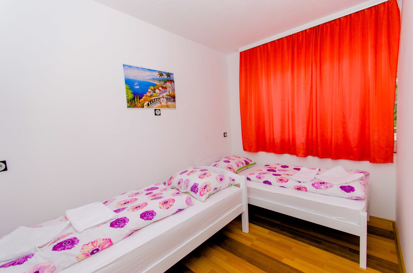 Ein kleines, sauberes Zimmer mit zwei Einzelbetten, einem roten Vorhang und einem Bild an der Wand.
