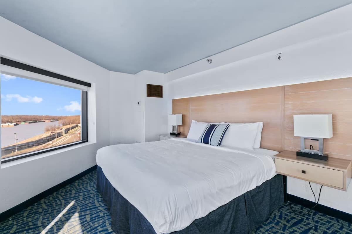 A hotel room with a large bed, two bedside tables, a window with a view of a body of water, and a wooden headboard.