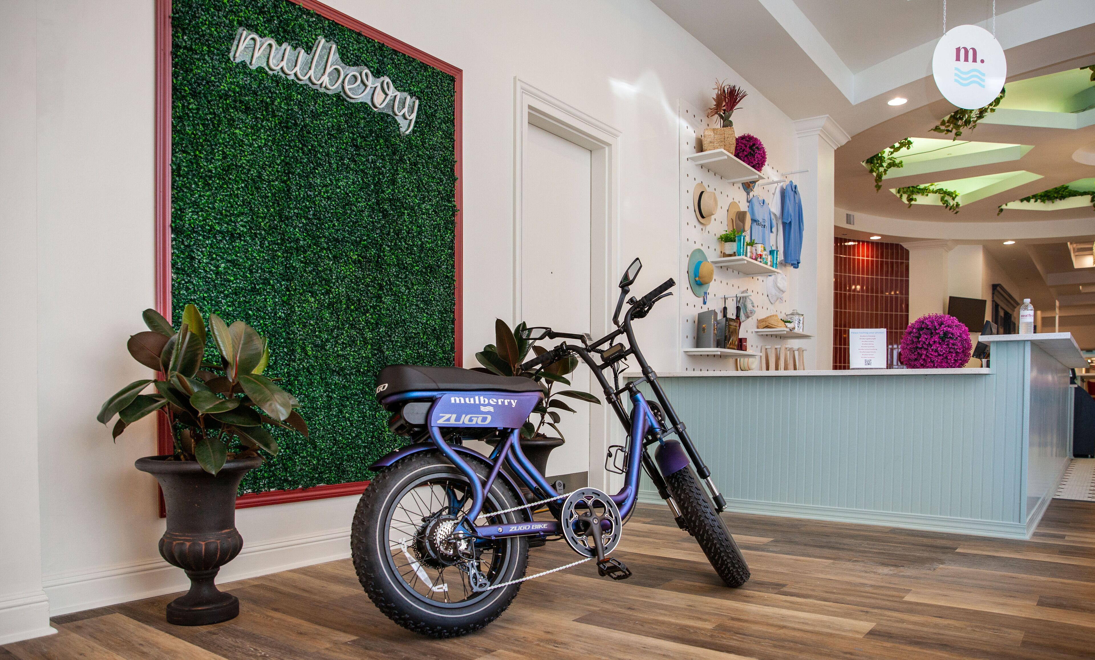 Een blauwe elektrische fiets met 'ZUGO'-branding, geparkeerd in een binnenruimte, een receptiebalie met een groene muur en 'mulberry'-aanduiding, en een potplant.
