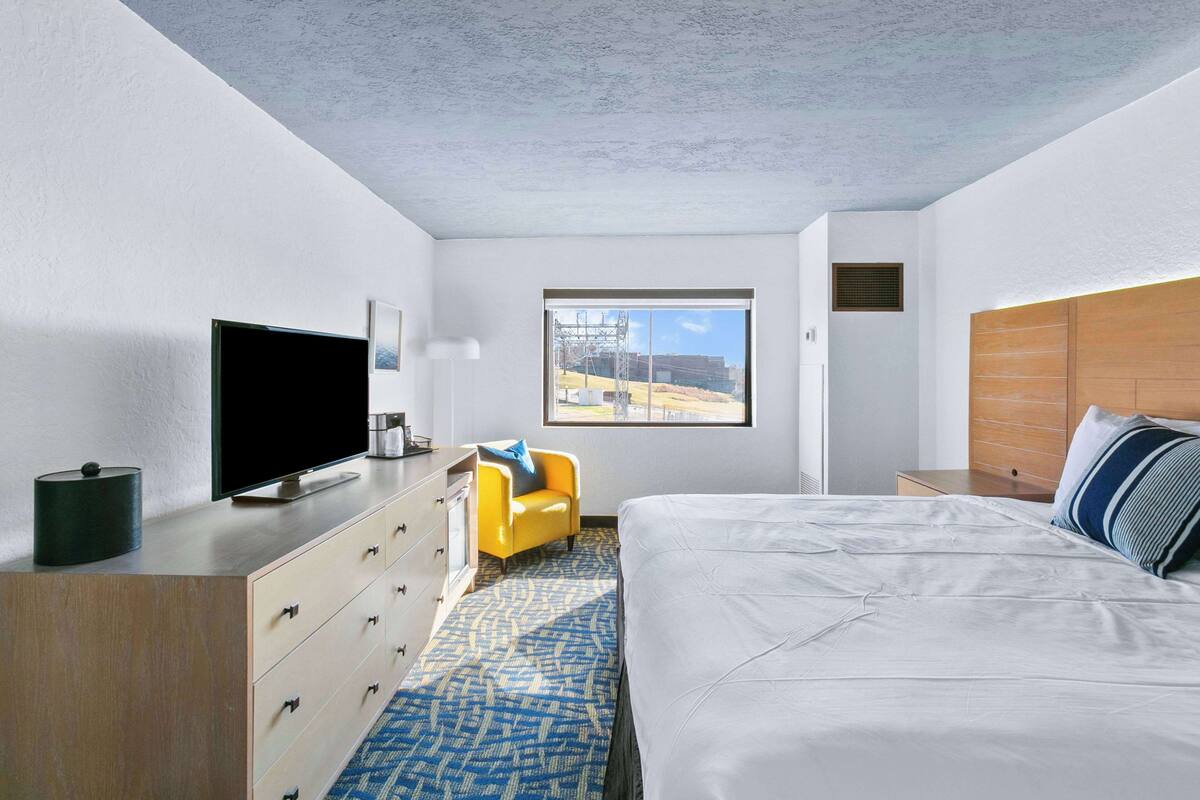 A modern hotel room with a large bed, a television, a desk with drawers, a yellow armchair, and a window with a view of the outdoors.