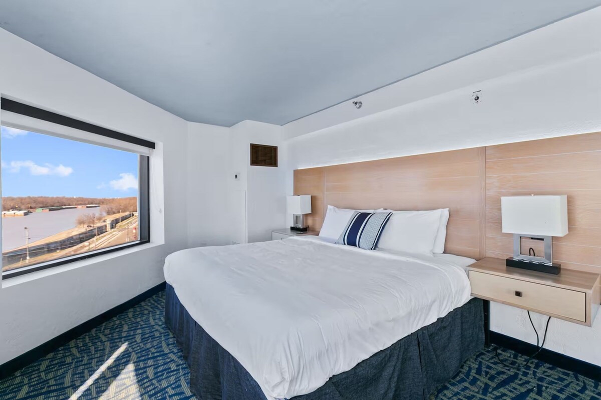 A hotel room with a large bed, two bedside tables, a window with a view of a body of water, and a wooden headboard.