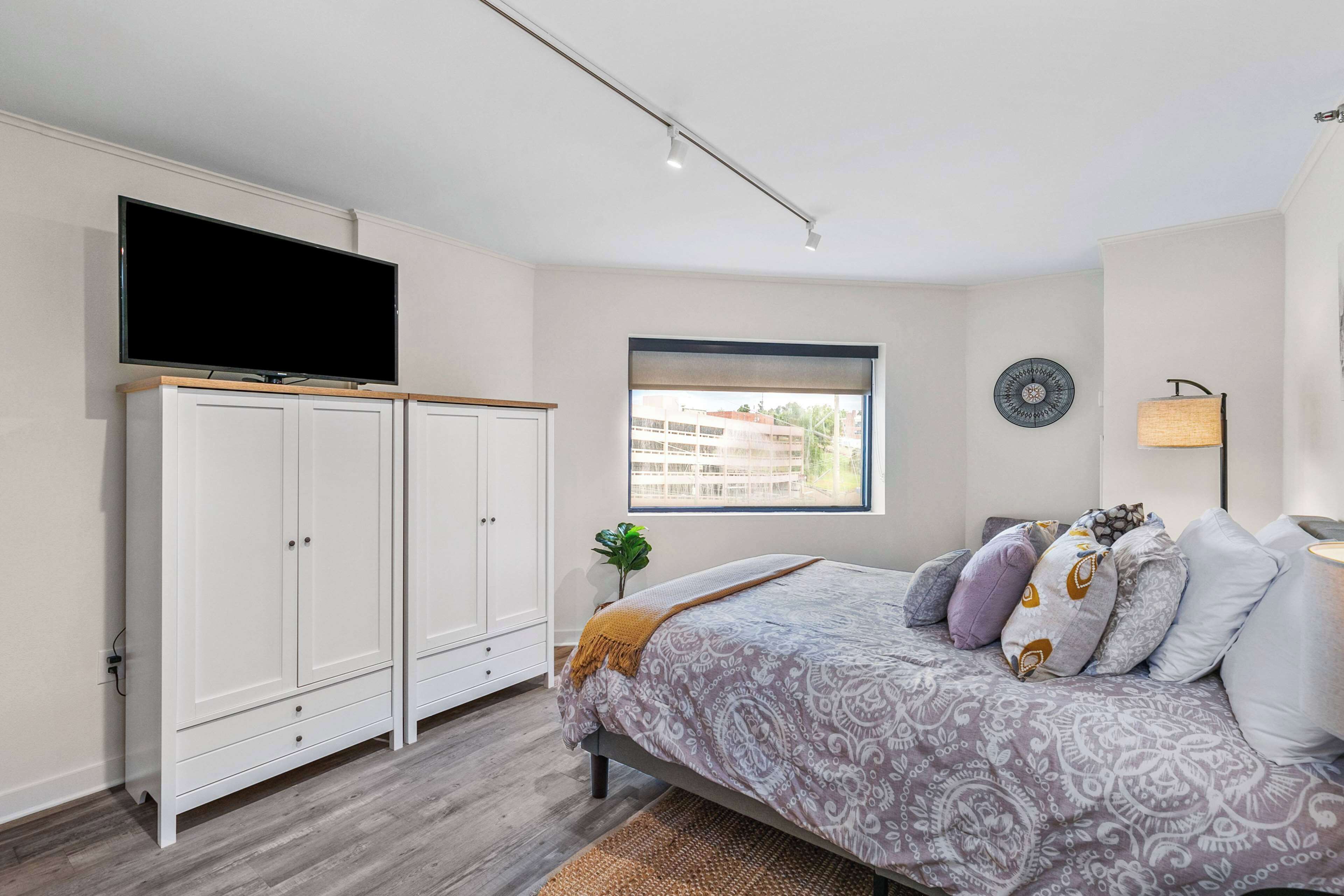 A bedroom with a bed, a TV mounted on a cabinet, a window, and a lamp.
