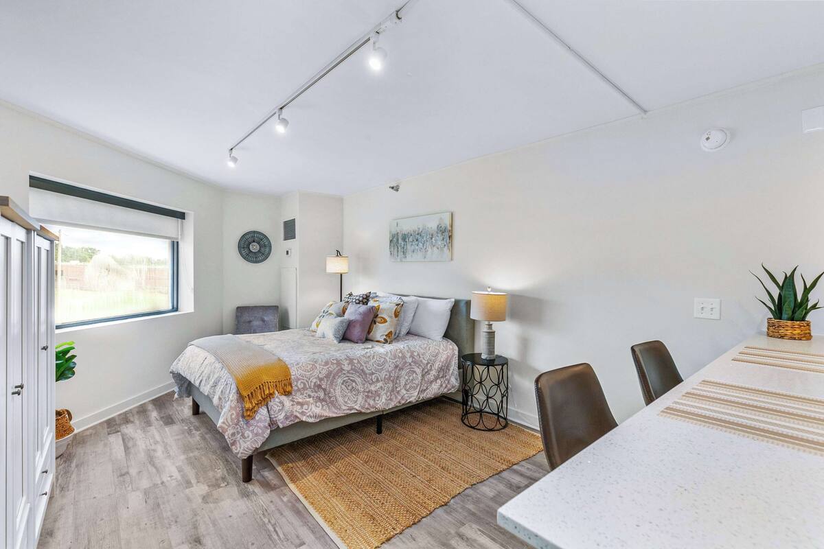 A bedroom with a bed, bedside table, lamp, and a view of the outside.