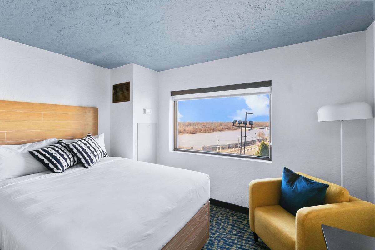 A hotel room with a bed, a window with a view, a yellow armchair, and a lamp.