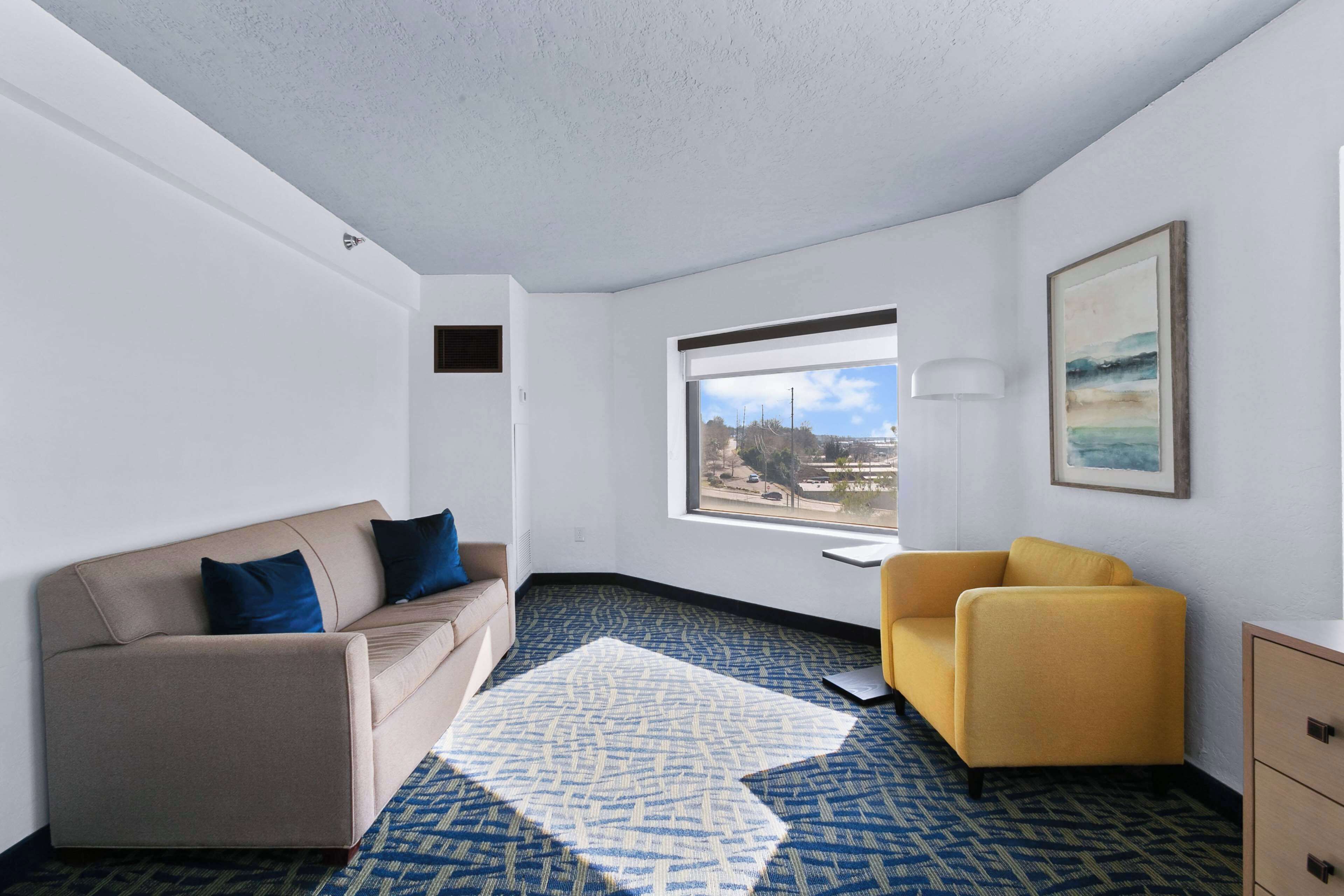 Habitación de hotel moderna con sofá color beige, un sillón amarillo, alfombra estampada, un ventanal con vista a la ciudad y un cuadro en la pared.