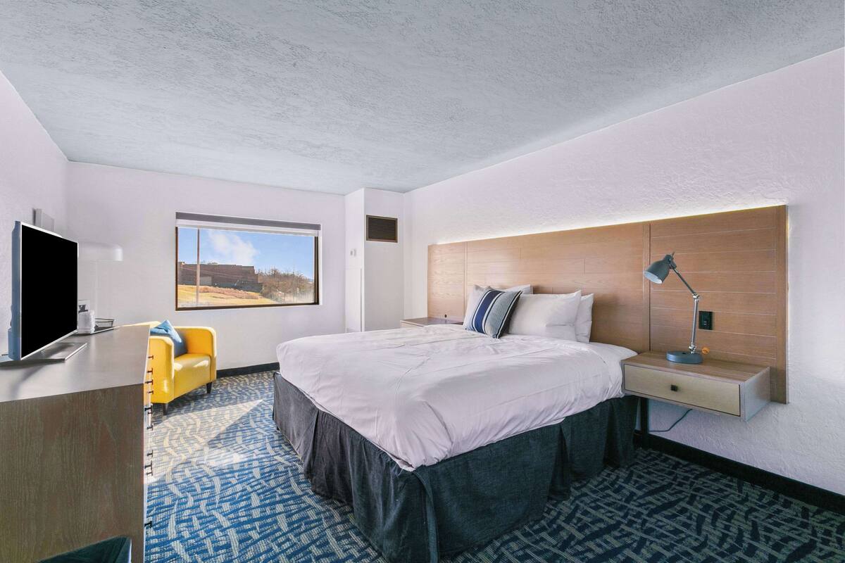 A modern hotel room with a large bed, a TV, a desk, and a window with a view of the outdoors.