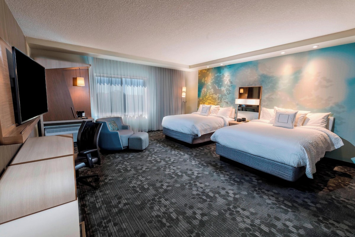 A hotel room with two beds, a desk, a TV, and a large window with curtains.