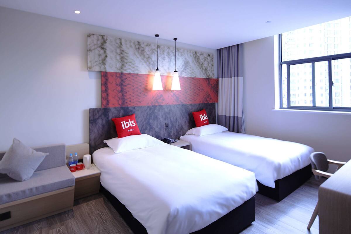 A hotel room with two beds, a sofa, a desk, and a window with a city view.