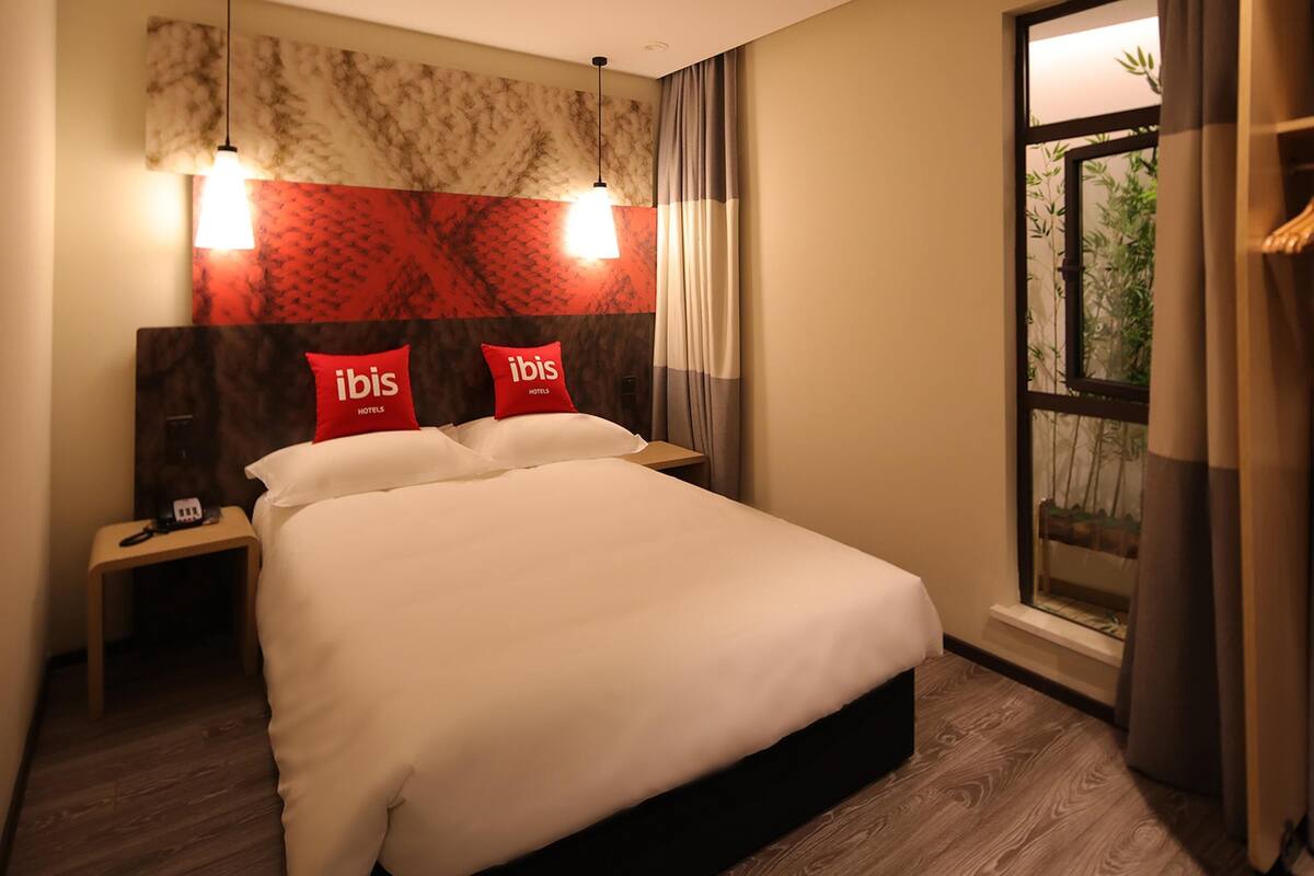 A hotel room with a large bed, two bedside tables, a nightstand, and a window with a view of greenery.