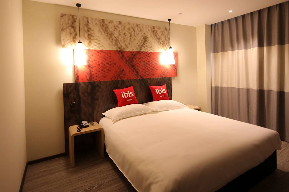 A hotel room with a large bed, two bedside tables, a nightstand, a telephone, and a headboard with 'ibis Hotels' branding.
