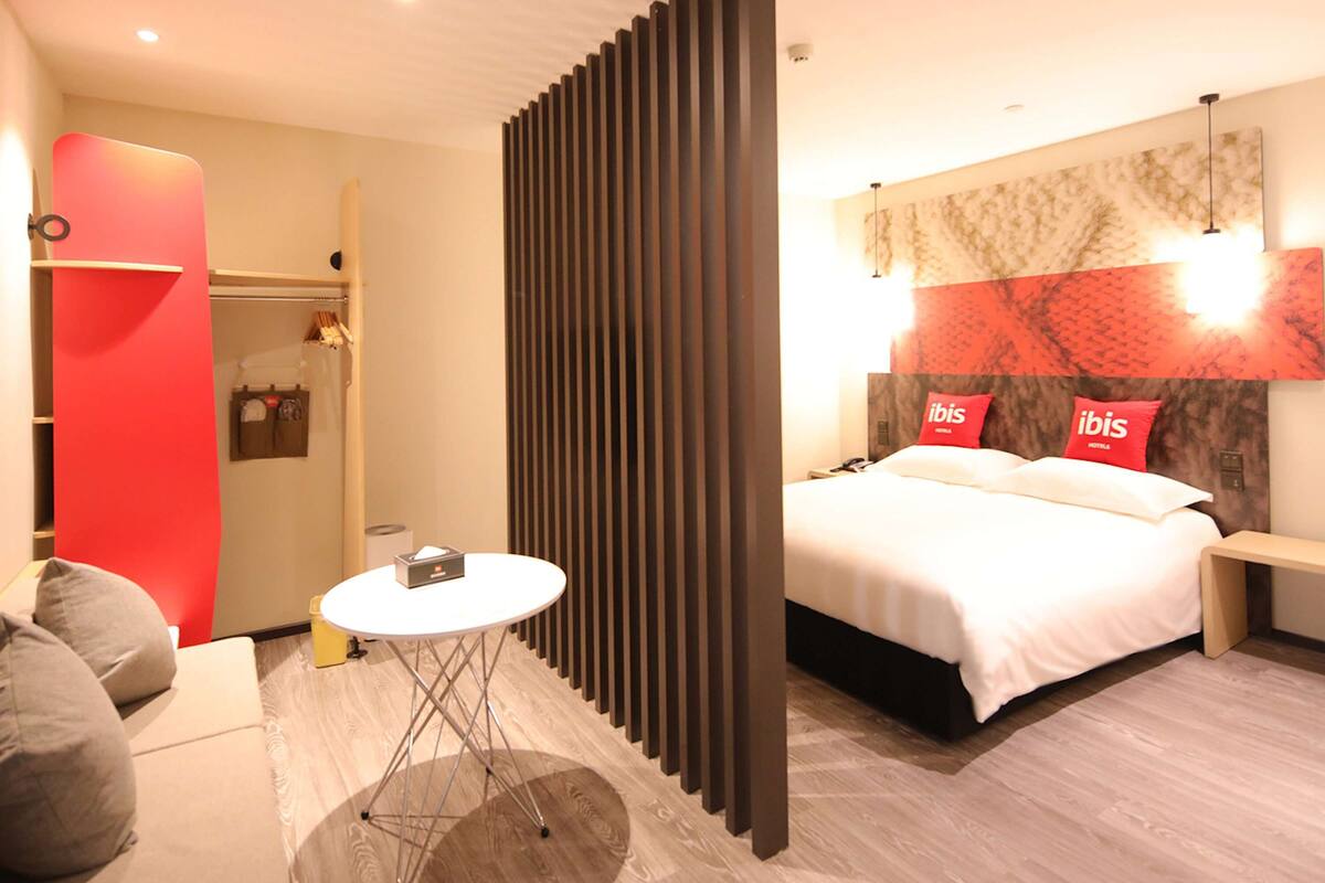 A modern hotel room with a bed, bedside table, and a small round table with a lamp.