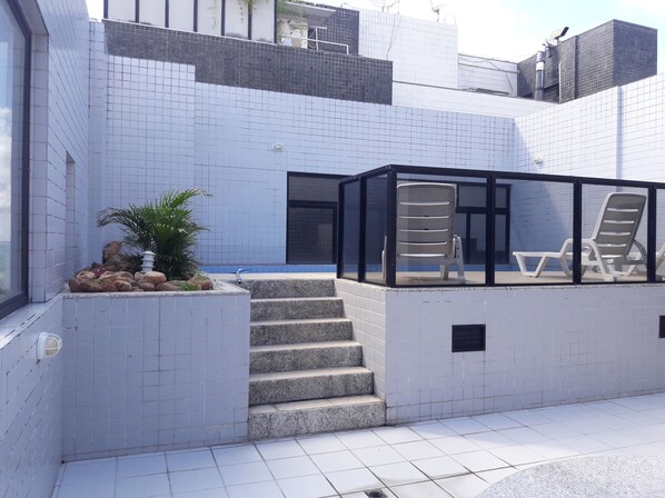 Un espace extérieur moderne avec des murs recouverts de carreaux, un escalier et un coin salon fermé par des parois de verre, équipé de fauteuils confortables.