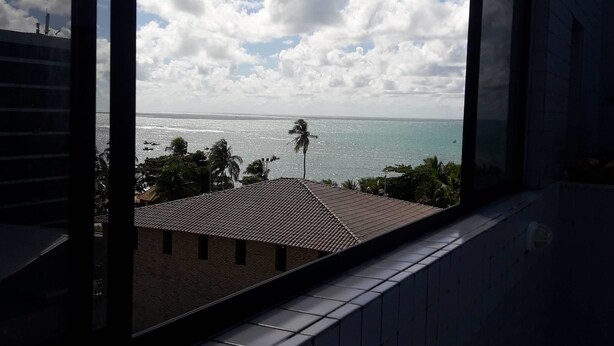 Vista da janela para uma praia, com céu claro e nuvens.