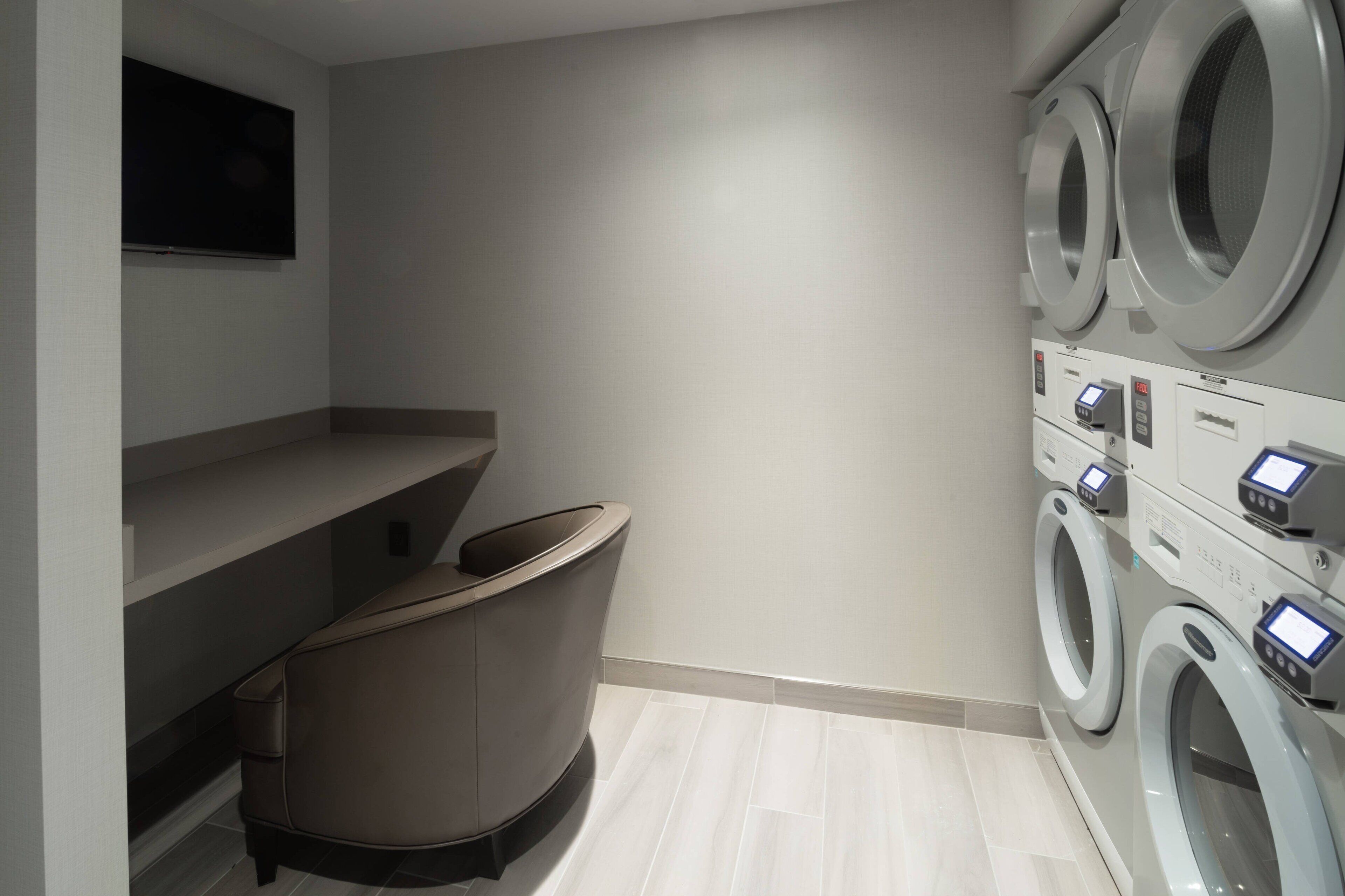 A modern laundry room with a built-in washer and dryer, a seating area, and a flat-screen TV mounted on the wall.