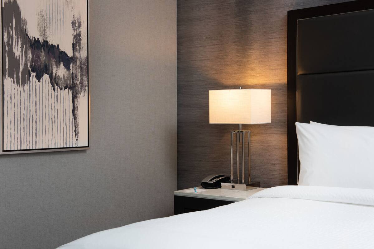 A hotel room with a bed, a bedside table with a lamp, a phone, and a painting on the wall.