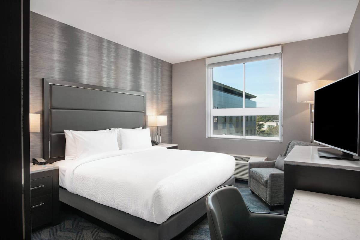 A modern hotel room with a large bed, a desk with a flat-screen TV, a chair, a desk lamp, and a window with a city view.