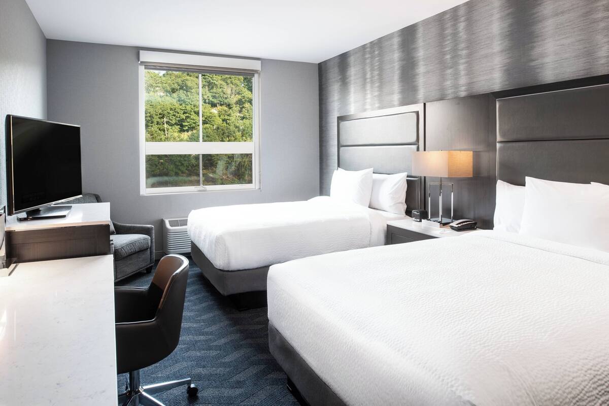 A hotel room with two beds, a desk with a flat-screen TV, a chair, and a window with a view of greenery.