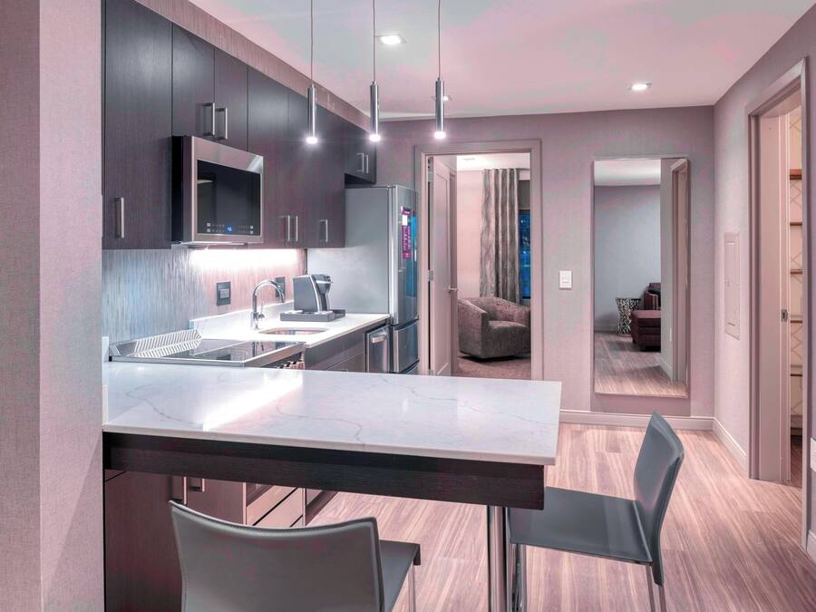 Cocina moderna con armarios oscuros, encimera de mármol y electrodomésticos de acero inoxidable. Cuenta con un comedor con mesa y sillas, y un pasillo que conduce a una sala de estar con sofá y mesa de centro.