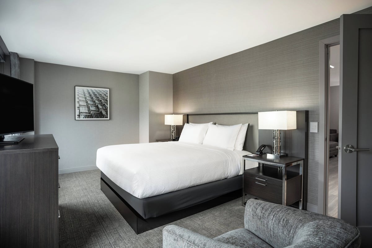 Een moderne hotelkamer met een groot bed, nachtkastjes, een flatscreen televisie en een grijze fauteuil.