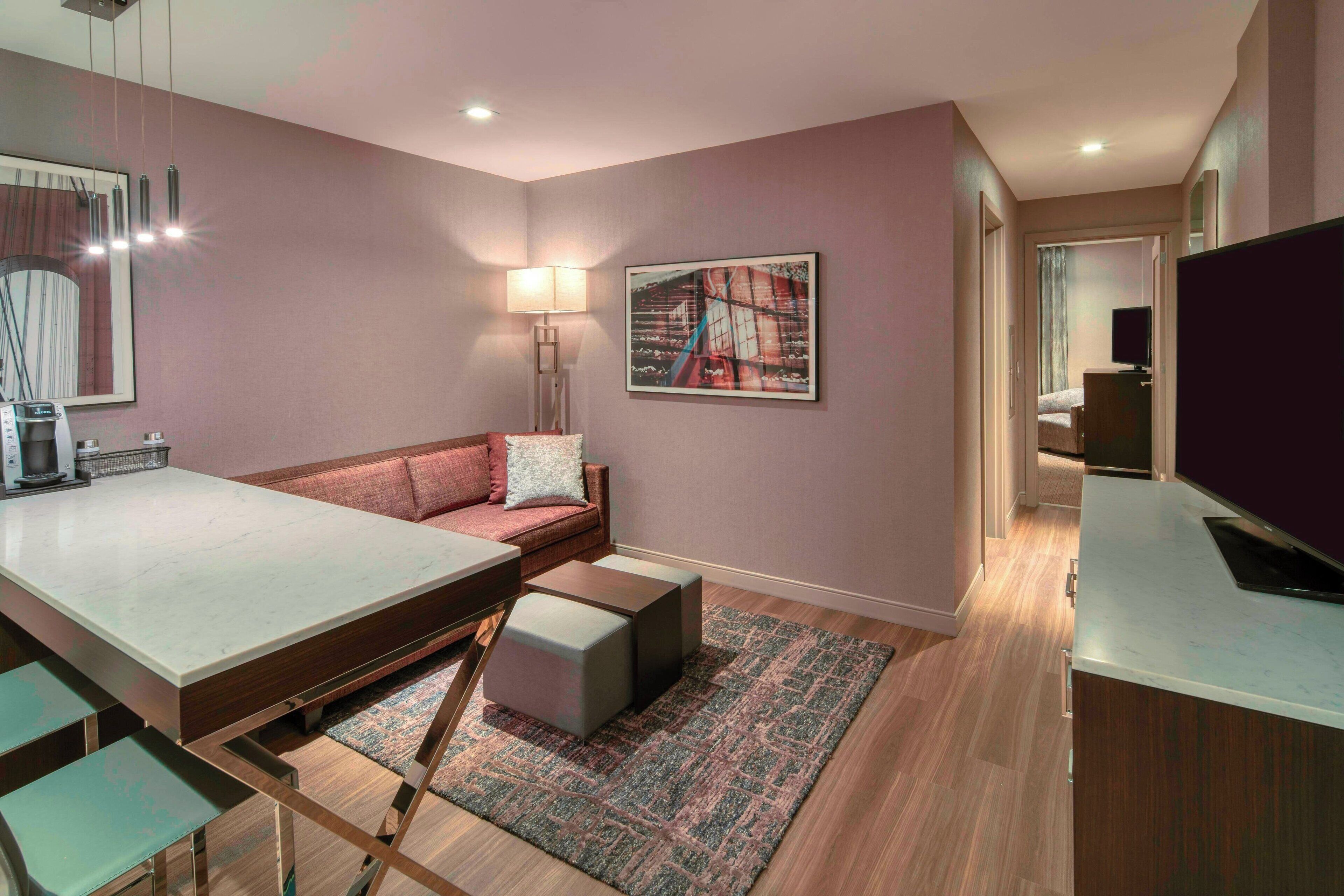 A modern hotel room with a sofa, a dining table, a TV, and a painting on the wall.