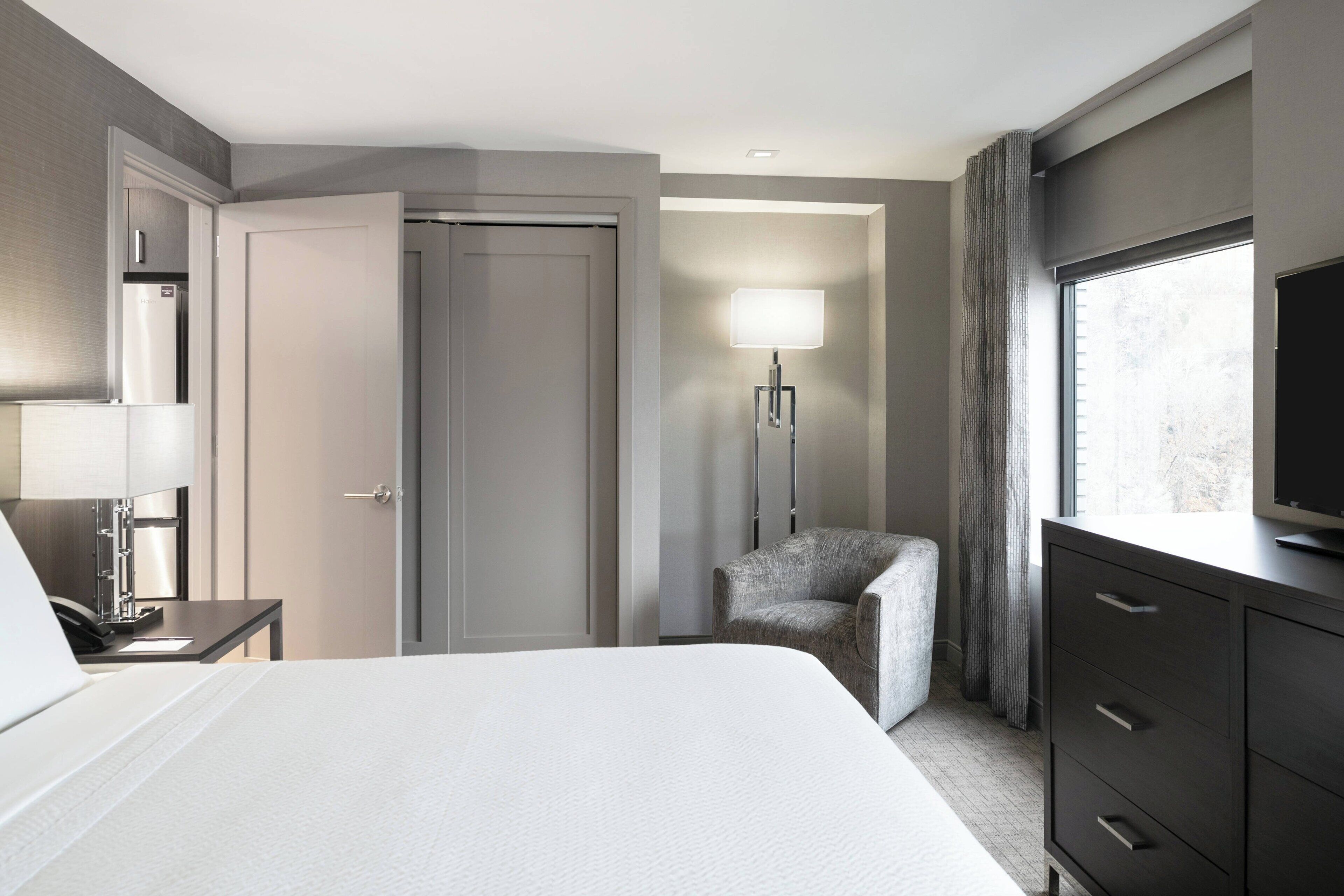 Ein Hotelzimmer mit einem Bett, einem Schreibtisch mit Lampe, einem Sessel, einem Kleiderschrank und einem Fernseher.