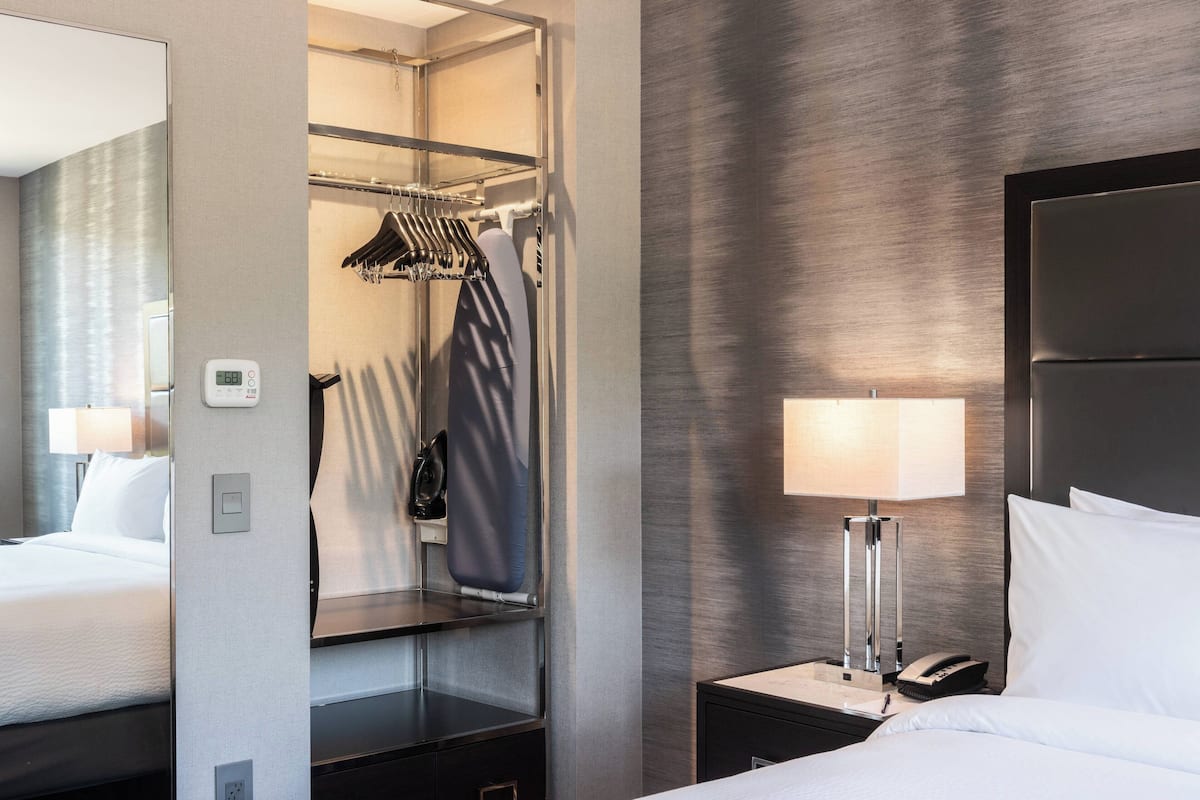 Ein Hotelzimmer mit Bett, Nachttisch mit Lampe, Spiegel, Kleiderschrank mit Kleidung und Bedienfeld an der Wand.